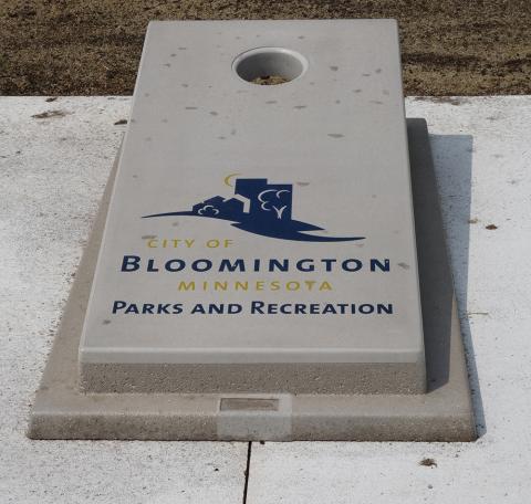 Gray concrete cornhole board with City of Bloomington Parks and Recreation logo in blue and gold