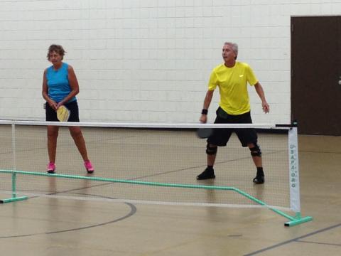 A woman in blue and man in yellow play pickleball indoors