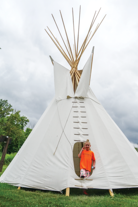 Child in an orange shirt emerging from a native American tipi. 