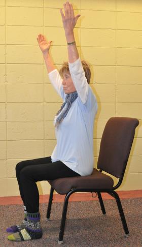 A woman with arms stretched over her head doing yoga on a chair.