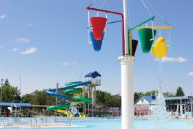Colorful image of red, blue, green and yellow bucket splash feature at Bloomington Family Aquatic Center with water slides and pool building in background