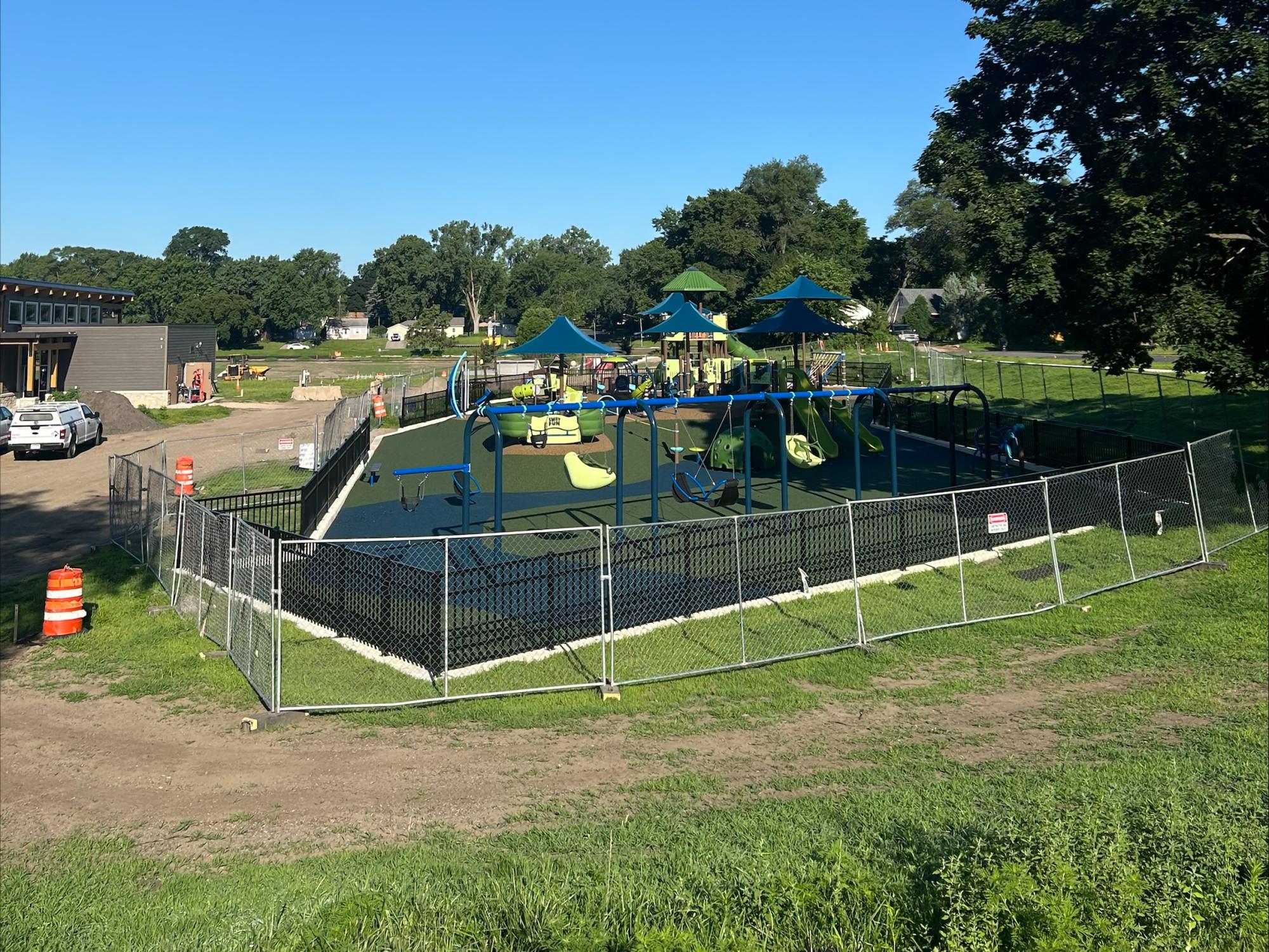 Bryant Park Construction July 8 2025: View of new inclusive playground with brightly colored blue and green apparatus including swings for younger children and new shelter building at left