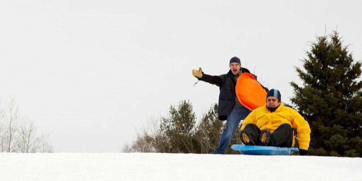 man in yellow coat on blue sled while man in black coat with orange sled cheers him on