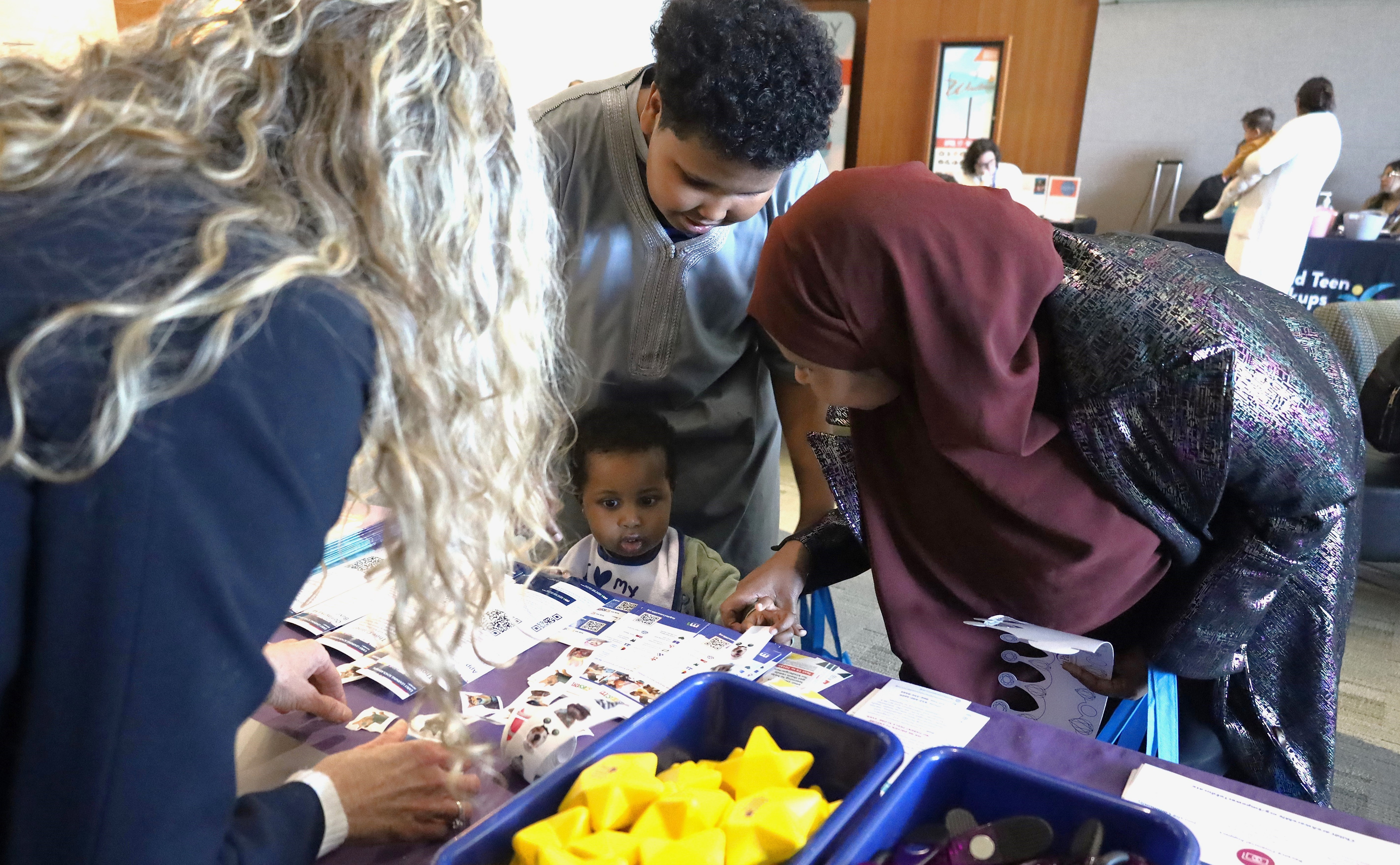 A Somali mom with her infant and older son standing in front of a table full of toys and resources talking to a lady behind the table. 