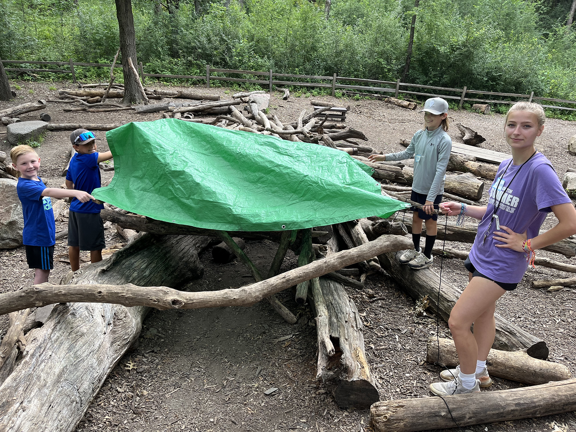Young people showing a survival shelter or "lean to" made from sticks and logs with a green tarp