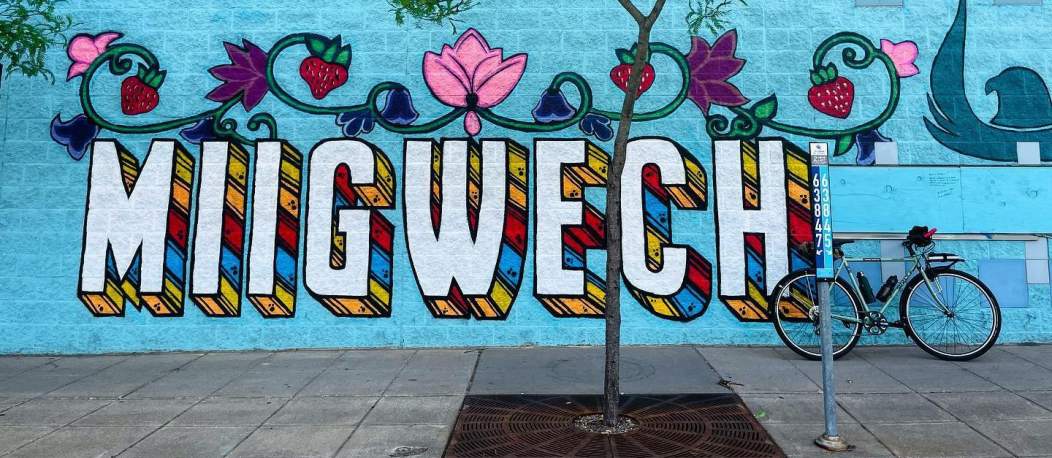 Mural by Thomazina TopBear, Holly Miskitoos Henning, and other community artists, blue wall with white text and flowers