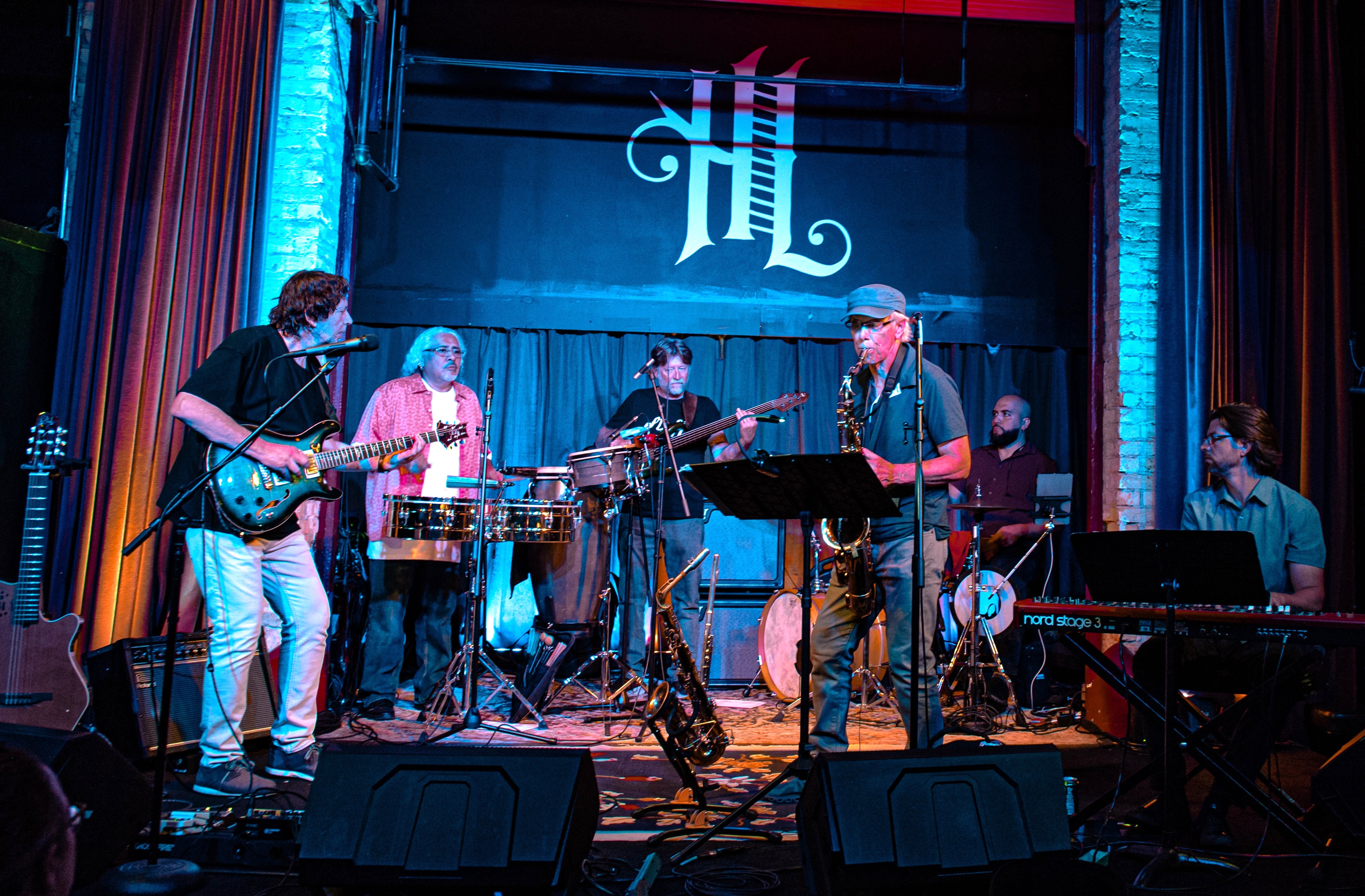 Band photo of Global Jazz Collegium playing guitars, drums, keyboard and saxophone on stage.