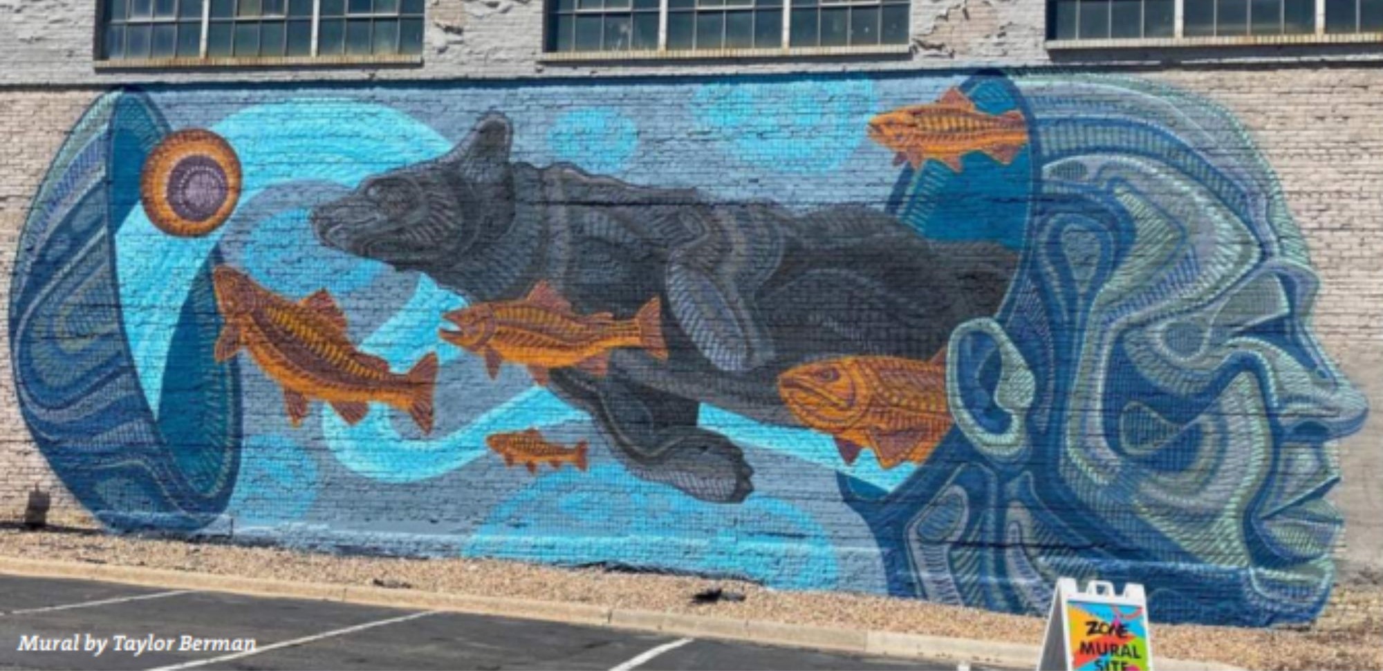 Photograph of a large outdoor mural painted on a brick wall. The mural shows a blue-toned face profile on the right, a cylindrical form on the left, several orange fish swimming across the scene, and a large dark animal figure in the center against swirling blue water-like shapes. A small sign near the lower right reads 'Zoe Mural Site'.