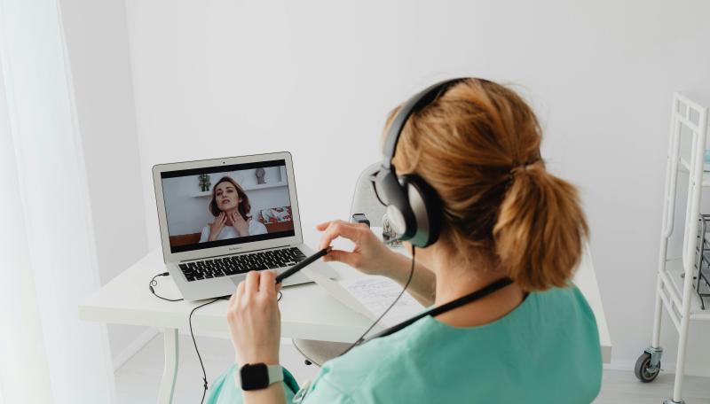 This is an image of a telehealth appointment or a virtual visit. A woman with dark, curly hair is seen on a screen touching both sides of her neck indicating she is not feeling well. Another woman is seen sitting and looking at the screen. She is the medical professional in sea foam-colored scrubs with her strawberry blonde hair pulled back into a pony tail. She is wearing headphones in order to hear her patient through the screen.  