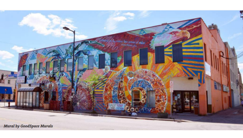 Photograph of a large, brightly colored mural covering the exterior walls of a corner building. The mural features geometric patterns, arches, birds, and a large eagle head near the roofline, with vivid orange, blue, red, yellow, and purple sections. A bus shelter stands on the sidewalk in front of the building.
