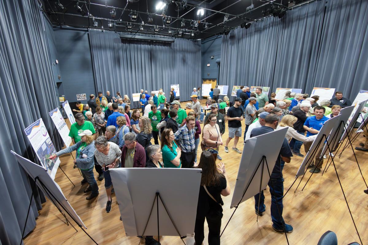 A large group of people are gathered indoors and looking at various display boards set up on easels.