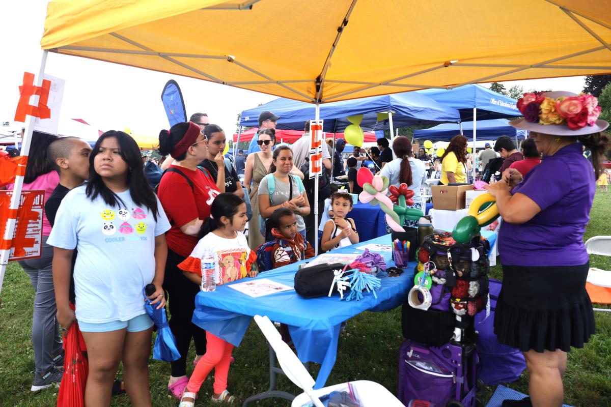 Under a large yellow tent, a group of adults and children gather around a vendor table at the Learning Linkup event.
