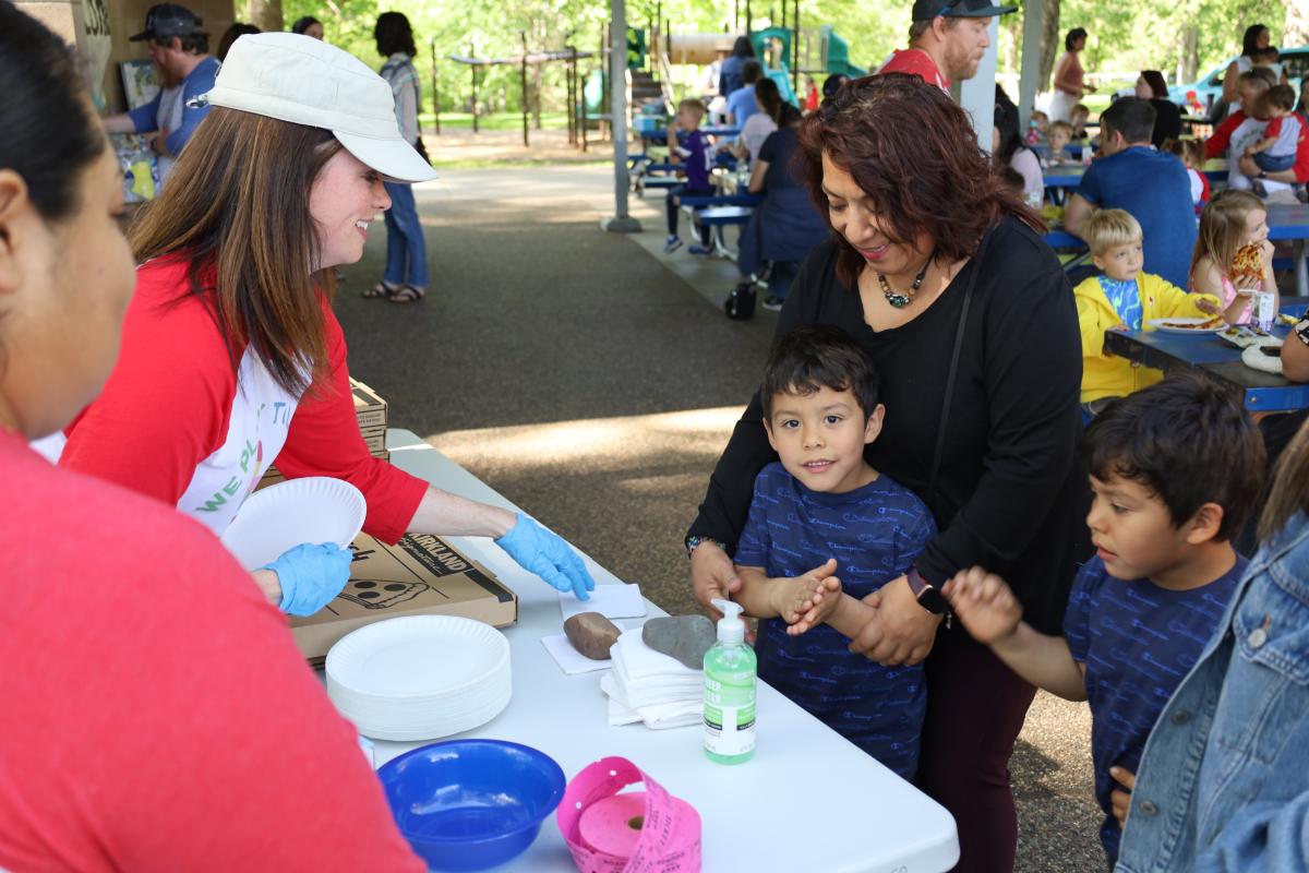 A woman in a white cap and red shirt, wearing blue gloves, smiles at another woman and two young boys standing at a table with paper plates and bowls.