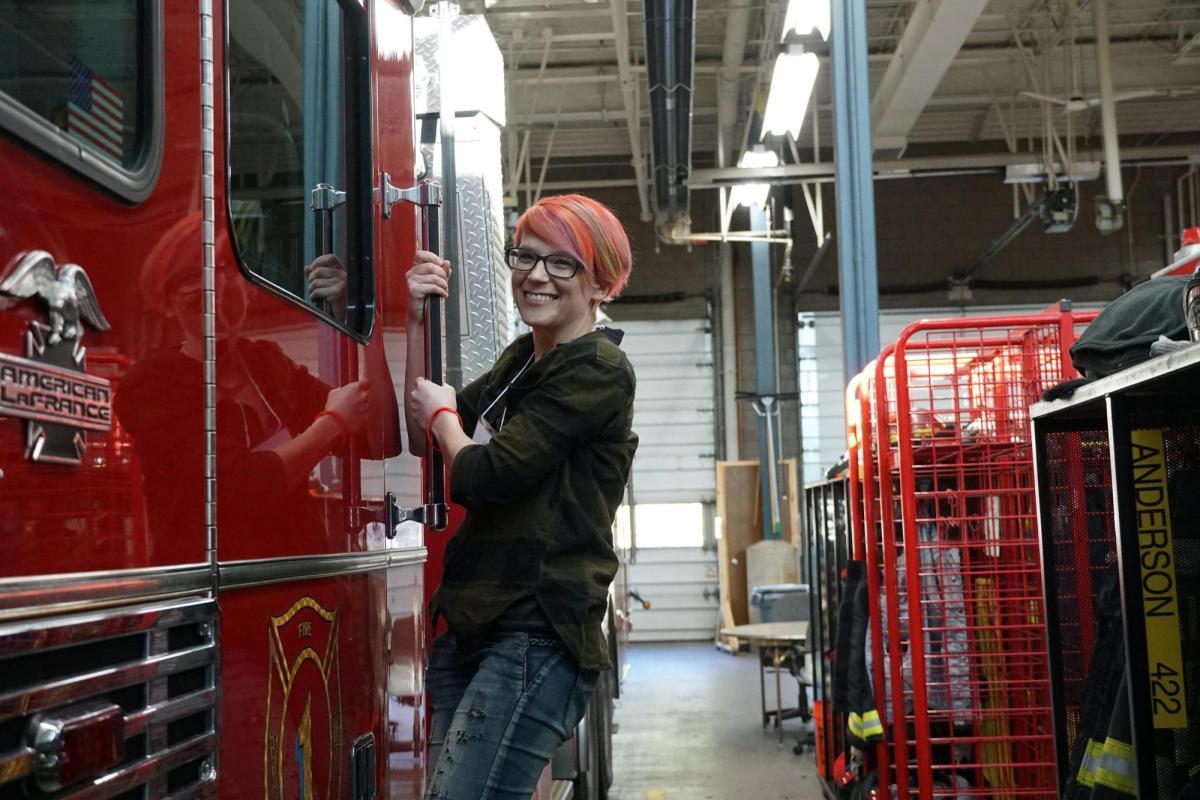 An adult holds onto the side of a firetruck while smiling at the camera