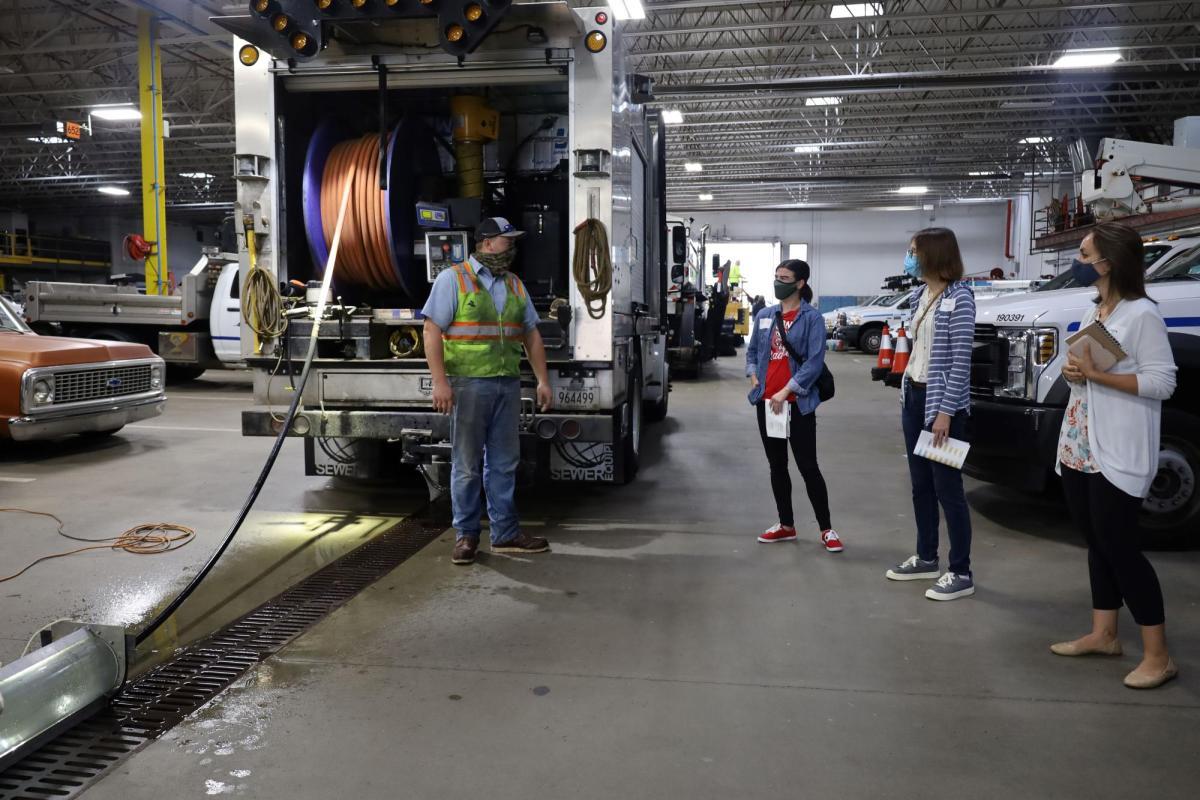 Three adults interact with a public works employee, standing in front of a vehicle with a large hose in the back