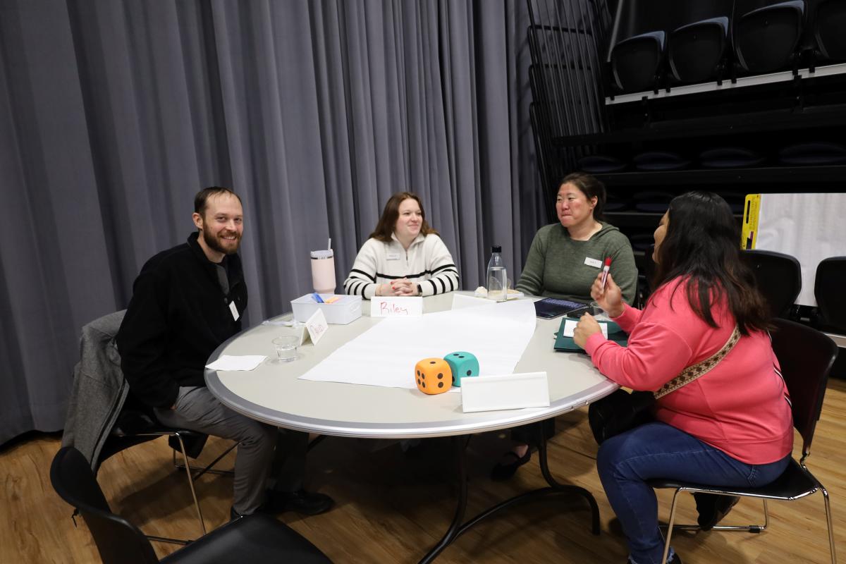A group of four adults sit at a table with paper, markers and large dice