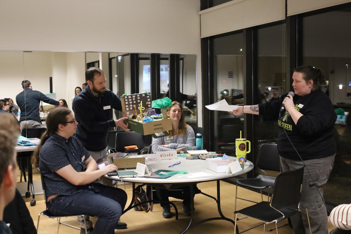 A person speaking into a microphone while presenting documents to a group seated around a table