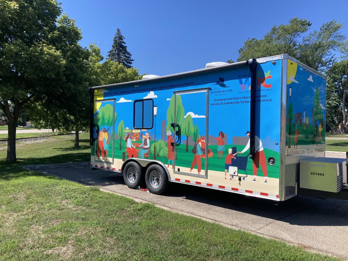 The image is of a ~20 foot trailer that has images of people of different races walking through a park on a trail: one person is using a cane, two others are embracing, two other are sitting on a bench enjoying coffee, one person is taking a photo of their friend in front of a tree, another person is pushing a stroller and has another child with them helping, an adult and a child are walking a dog, two people are running, one person has their arms raised in a stretch. There are stands in the park with coffe