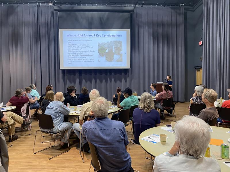 A room of ~40 people siting around circular tables looking forward at a presentation on a large screen.