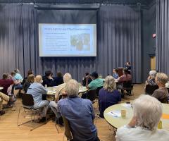 A room of ~40 people siting around circular tables looking forward at a presentation on a large screen.