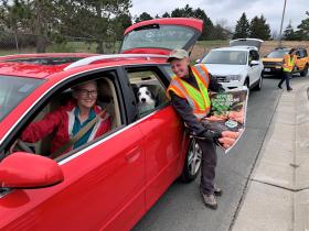 Person holding bag of compost next to a red car in the pickup line. Woman driving and dog looking out the window. All smiling at the camera.